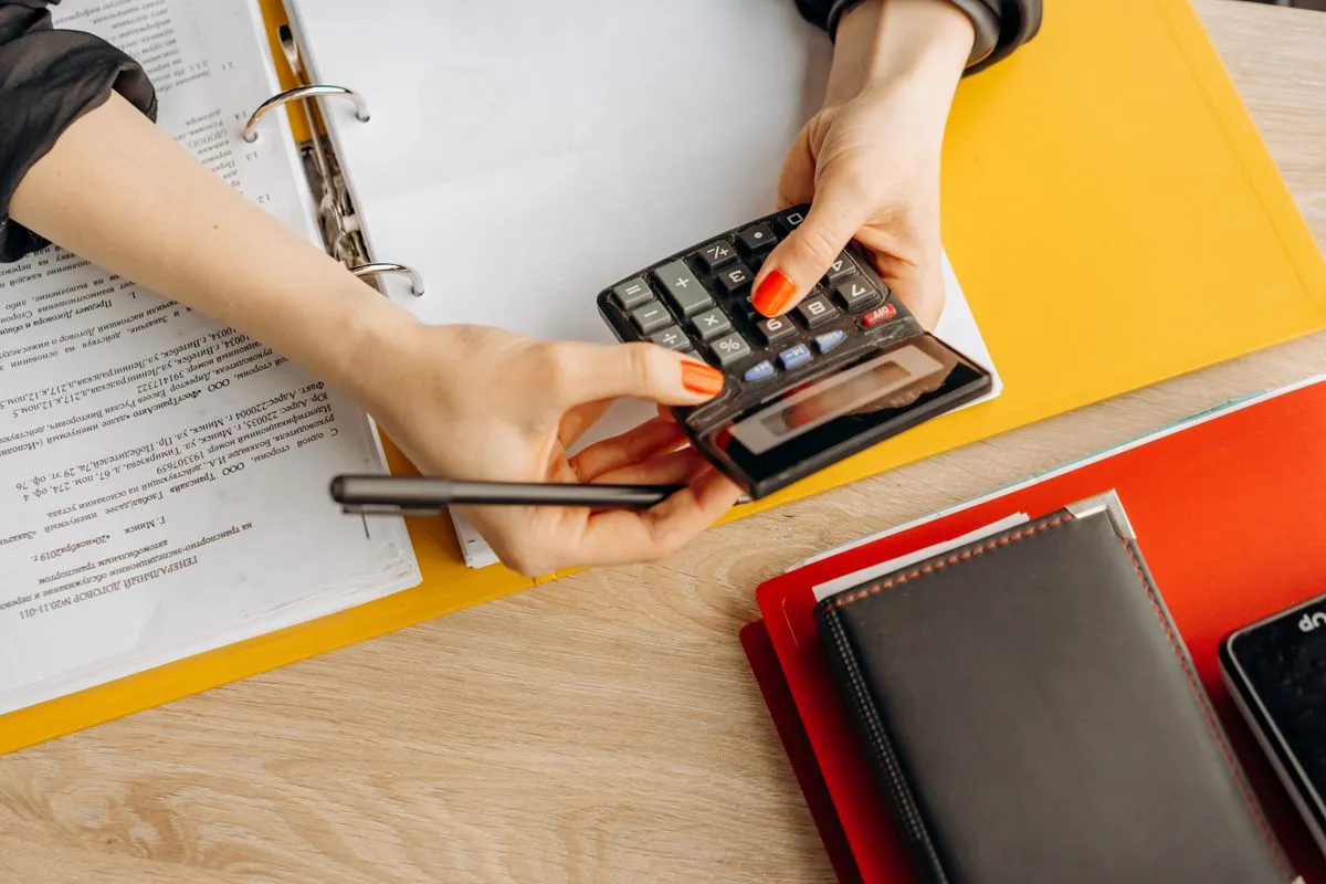 Mãos usando calculadora sobre uma mesa com documentos e pastas, organizando contas a pagar e a receber