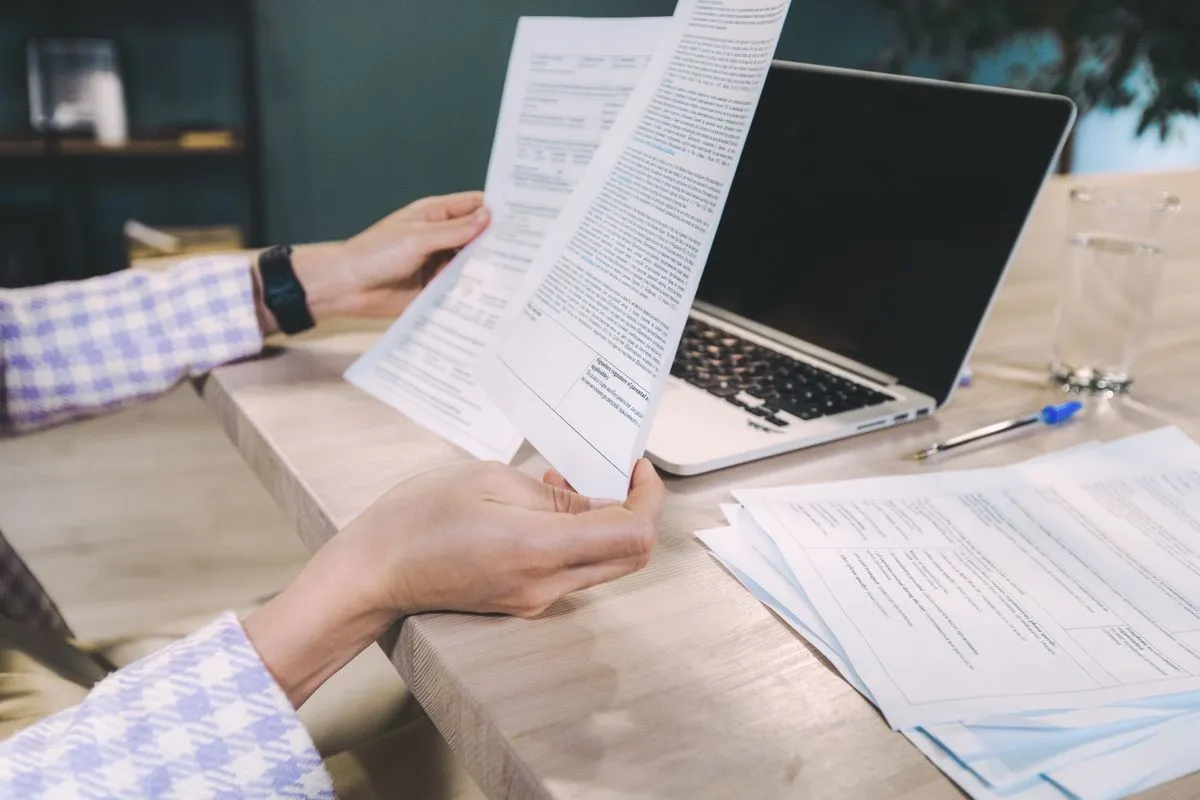 Mãos consultando documentos impressos ao lado de um notebook sobre a mesa