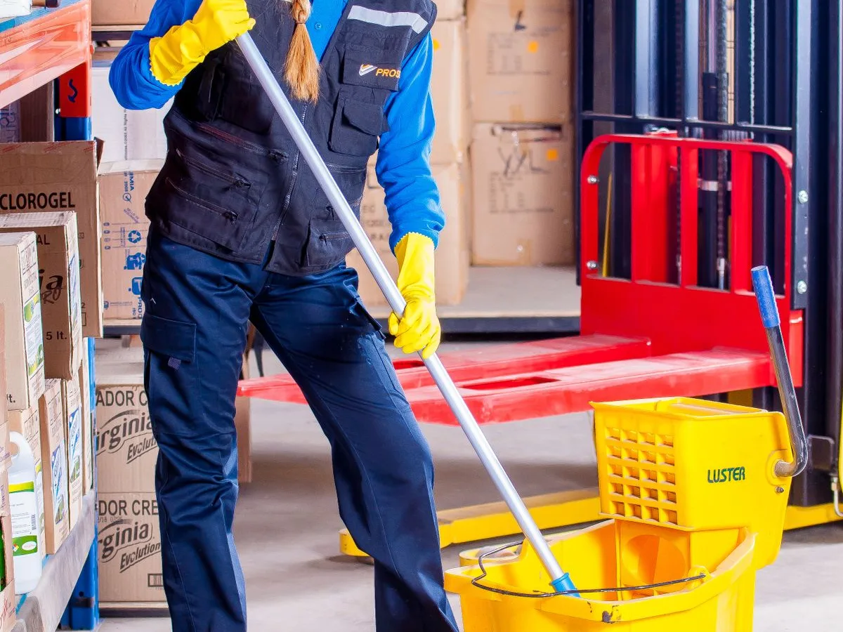 Funcionária de limpeza com equipamento de proteção passando rodo no piso de um galpão
