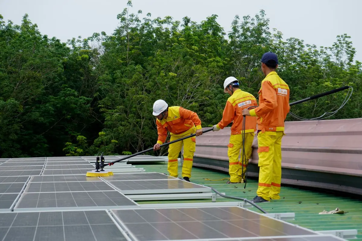 Três técnicos uniformizados e de capacete limpando módulos solares no telhado